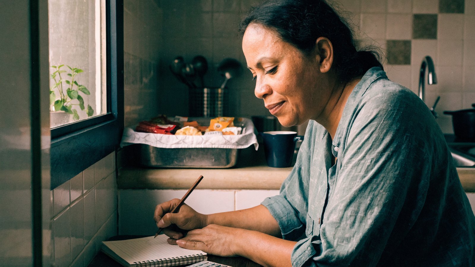 Mulher sentada à mesa da cozinha escrevendo em um caderno com calculadora ao lado, com a produção pronta ao fundo.