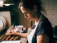 Mulher de avental em cozinha doméstica anotando em um caderno ao lado de uma bandeja de docinhos, com expressão cansada.