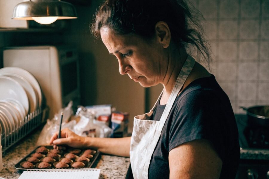 Mulher de avental em cozinha doméstica anotando em um caderno ao lado de uma bandeja de docinhos, com expressão cansada.
